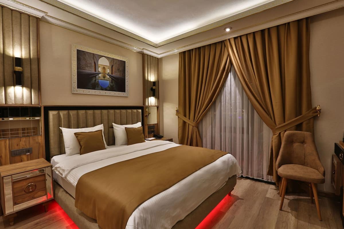 A hotel room with a large bed, a wooden nightstand, a chair, and a large window with curtains.