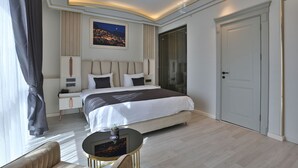 A modern hotel room with a large bed, a bedside table, a chair, a small round table, and a large window with curtains. - Anemon Mardin Otel (Kiziltepe)