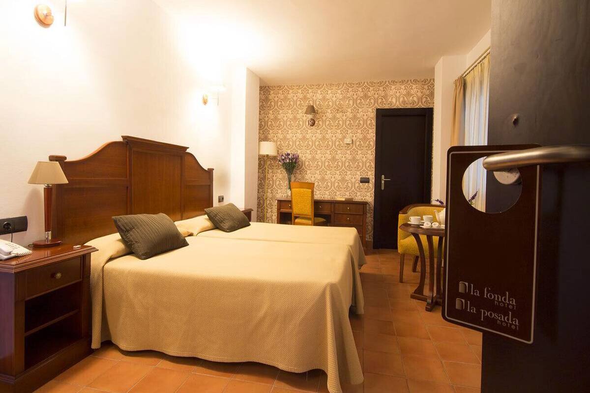 Habitación de hotel con dos camas, una mesita de noche con lámpara, una pequeña mesa con silla y un adorno en la pared.