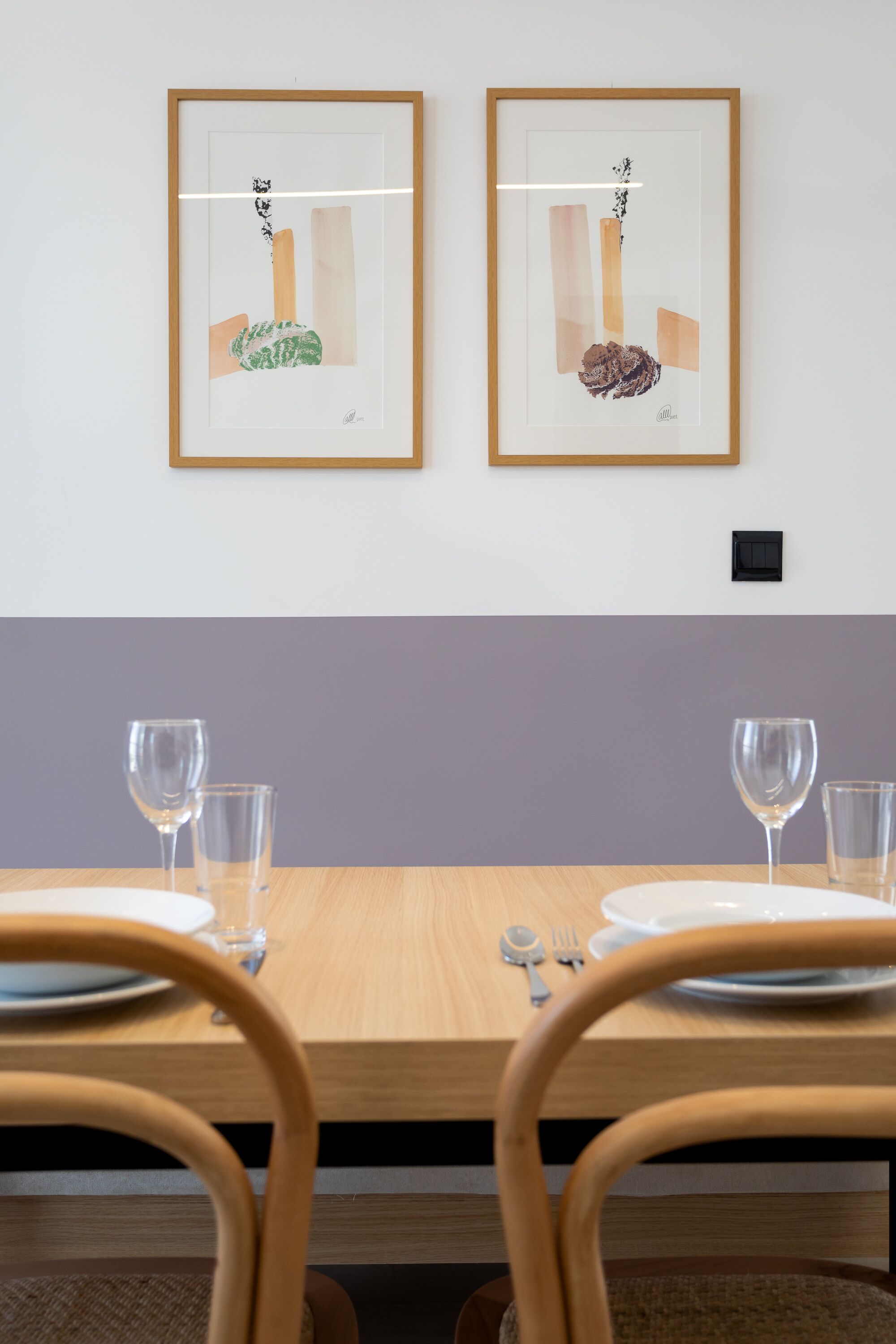 A dining table with two framed artworks, a light purple wall, and a wooden chair in the foreground.