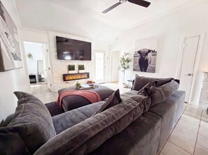 A modern living room with a grey sofa, a flat-screen TV mounted on the wall, a fireplace, and a ceiling fan. - Luxury Modern 4BR | Hot Tub | Mins to DT & Rainey (Austin)