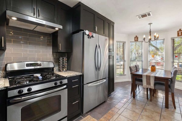 A modern kitchen with dark cabinetry, stainless steel appliances, and a dining area with a table and chairs. - Luxury Modern 4BR | Hot Tub | Mins to DT & Rainey (Austin)
