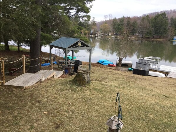A lakeside gazebo with a small deck, surrounded by a rope fence, near a dock with boats.