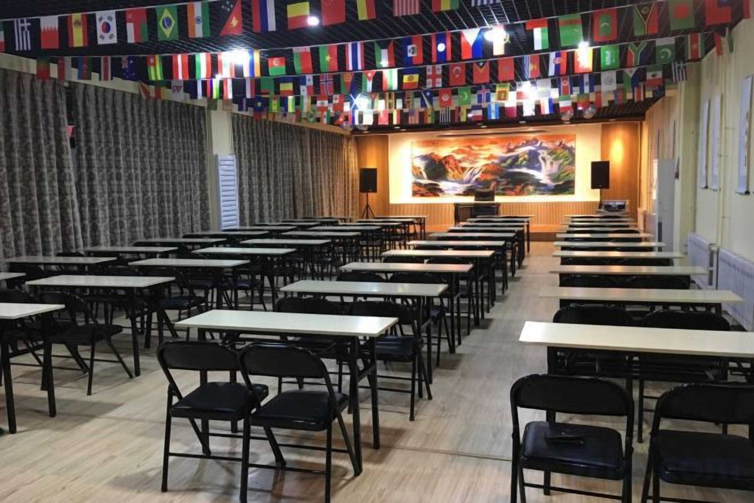 A conference room with rows of tables and chairs, a podium, and a backdrop with flags.