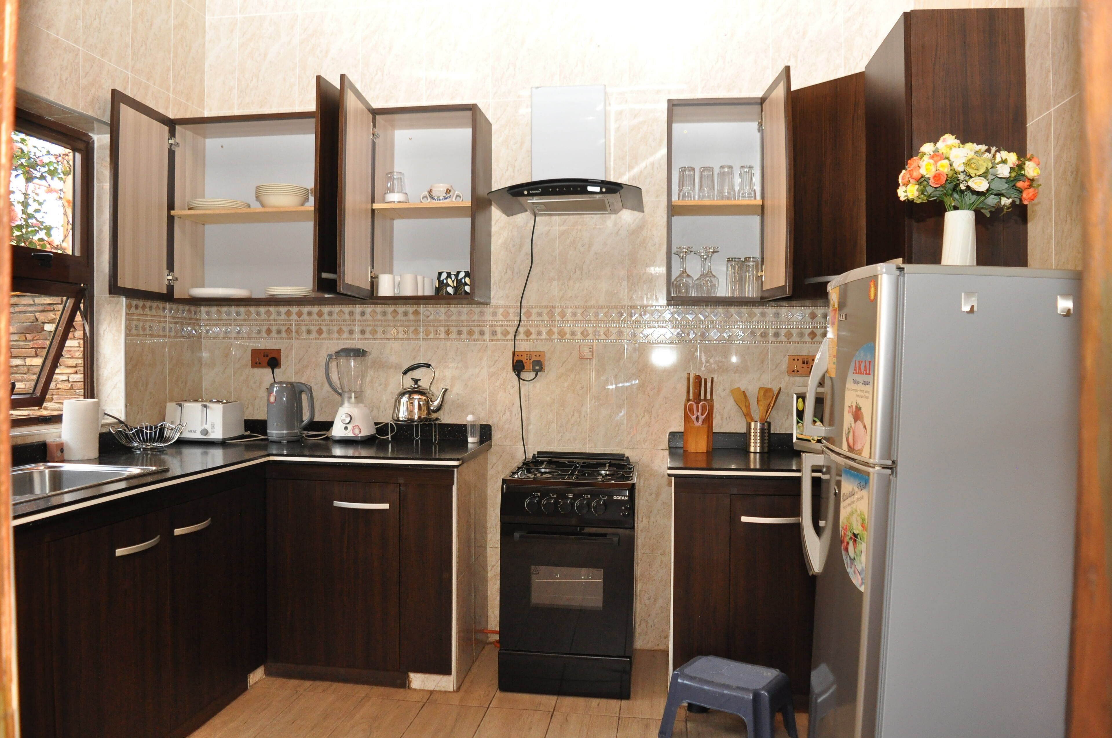Cocina compacta con armarios de madera oscura, estufa a gas, horno integrado, refrigerador y un taburete pequeño.