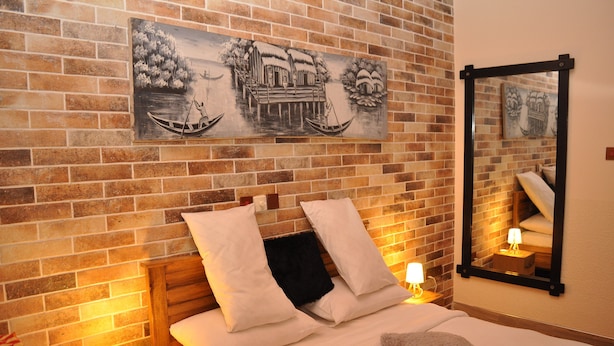 Een hotelkamer met een bakstenen muur, een bed met witte beddengoed, twee kussens, een nachtkastje met een lamp, en een schilderij van een traditioneel tafereel.