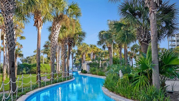 A curved swimming pool surrounded by palm trees and greenery. - Destin West Gulfside 301 ~ True Beachfront Luxury ~ Beautiful Views! (Fort Walton Beach)