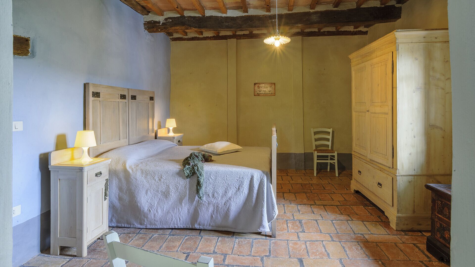 A bedroom with a bed, bedside tables, a chair, a wardrobe, and a hanging light fixture.