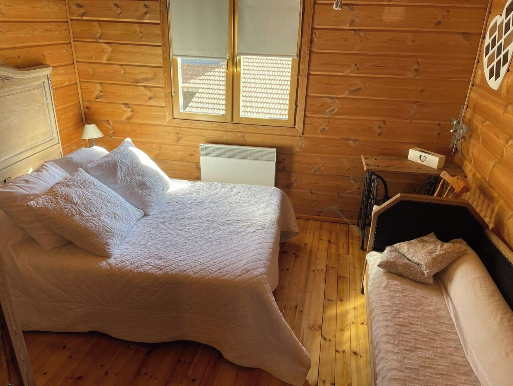 Une chambre dans une cabane en bois, avec un lit, une petite table de chevet, une chaise et une fenêtre avec des stores.