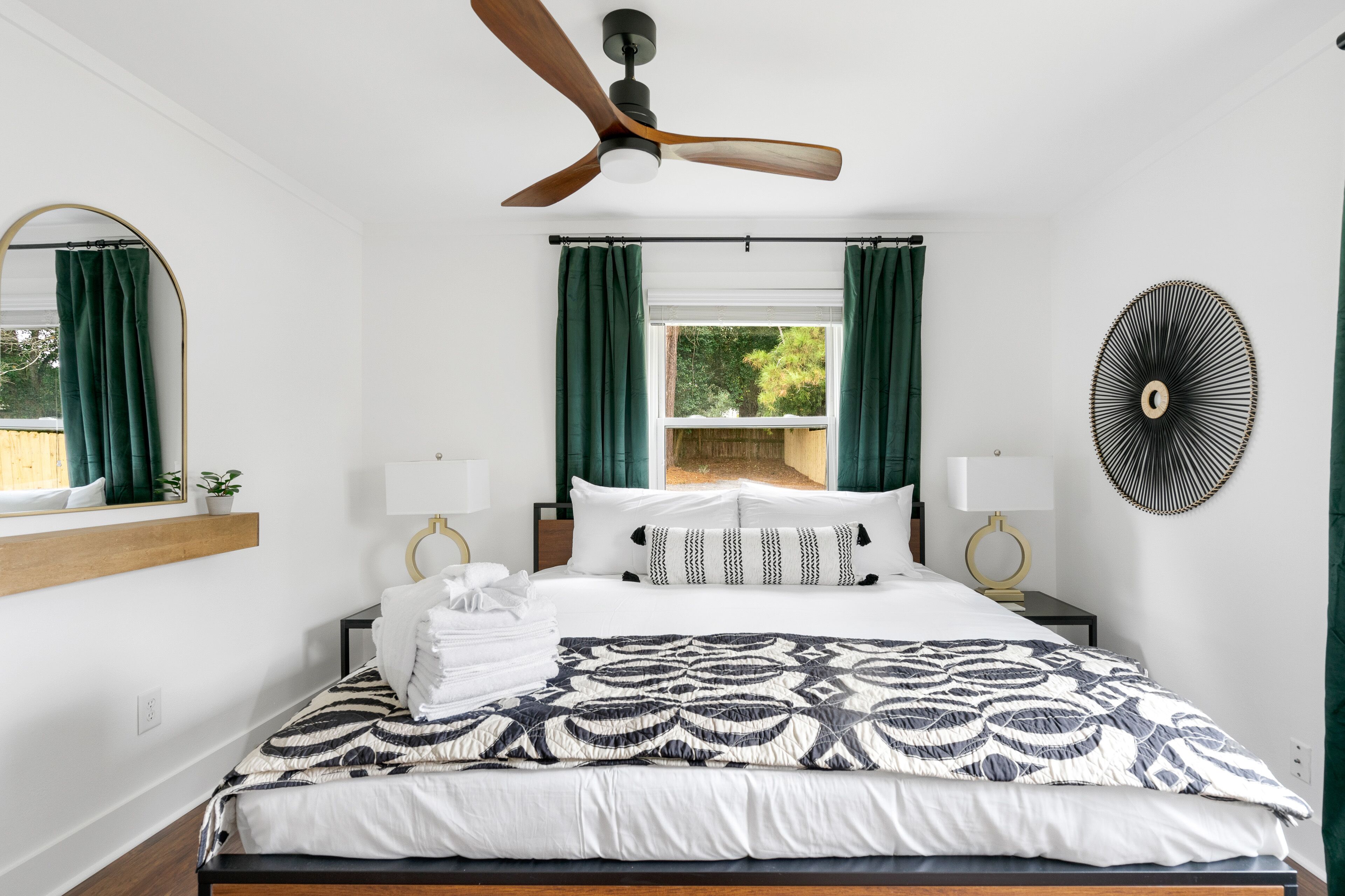 A neatly arranged bedroom with a bed, a ceiling fan, a window with curtains, a wall-mounted fan, and a mirror.