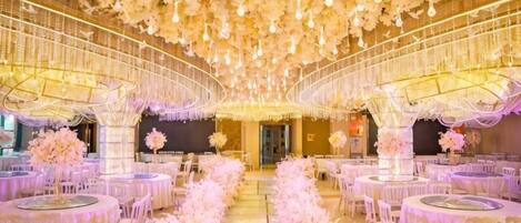 Un salón de banquetes con mesas redondas dispuestas en filas, un pasillo central y un techo decorado con luces colgantes y arreglos florales.