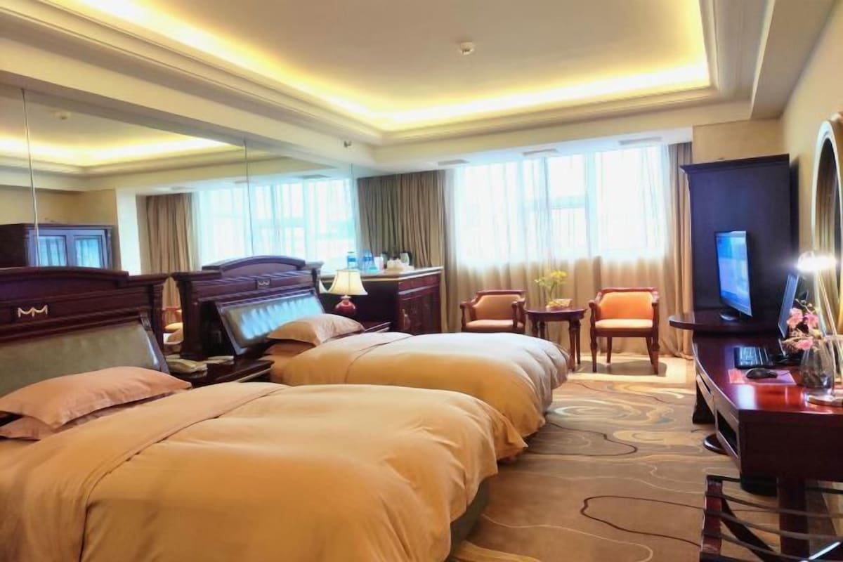 A hotel room with two beds, a desk, a TV, and a large window with curtains.