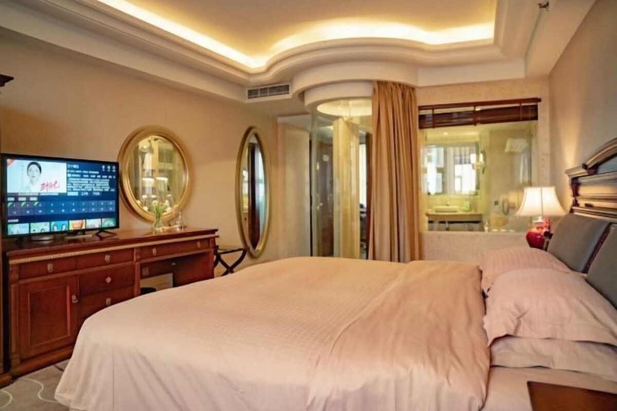 A hotel room with a large bed, a television on a wooden dresser, a mirror, and a window with curtains.