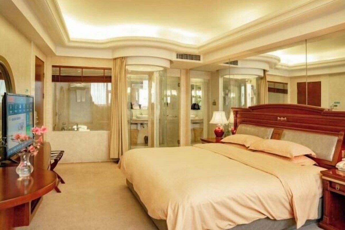 A hotel room with a large bed, a desk with a computer, a mirror, and a bathroom with a glass door.