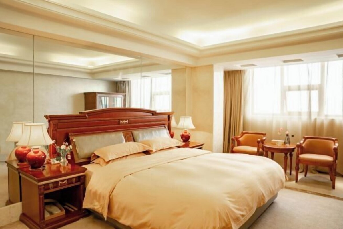 A hotel room with a large bed, bedside tables, lamps, a mirror, a wardrobe, and a seating area with chairs.