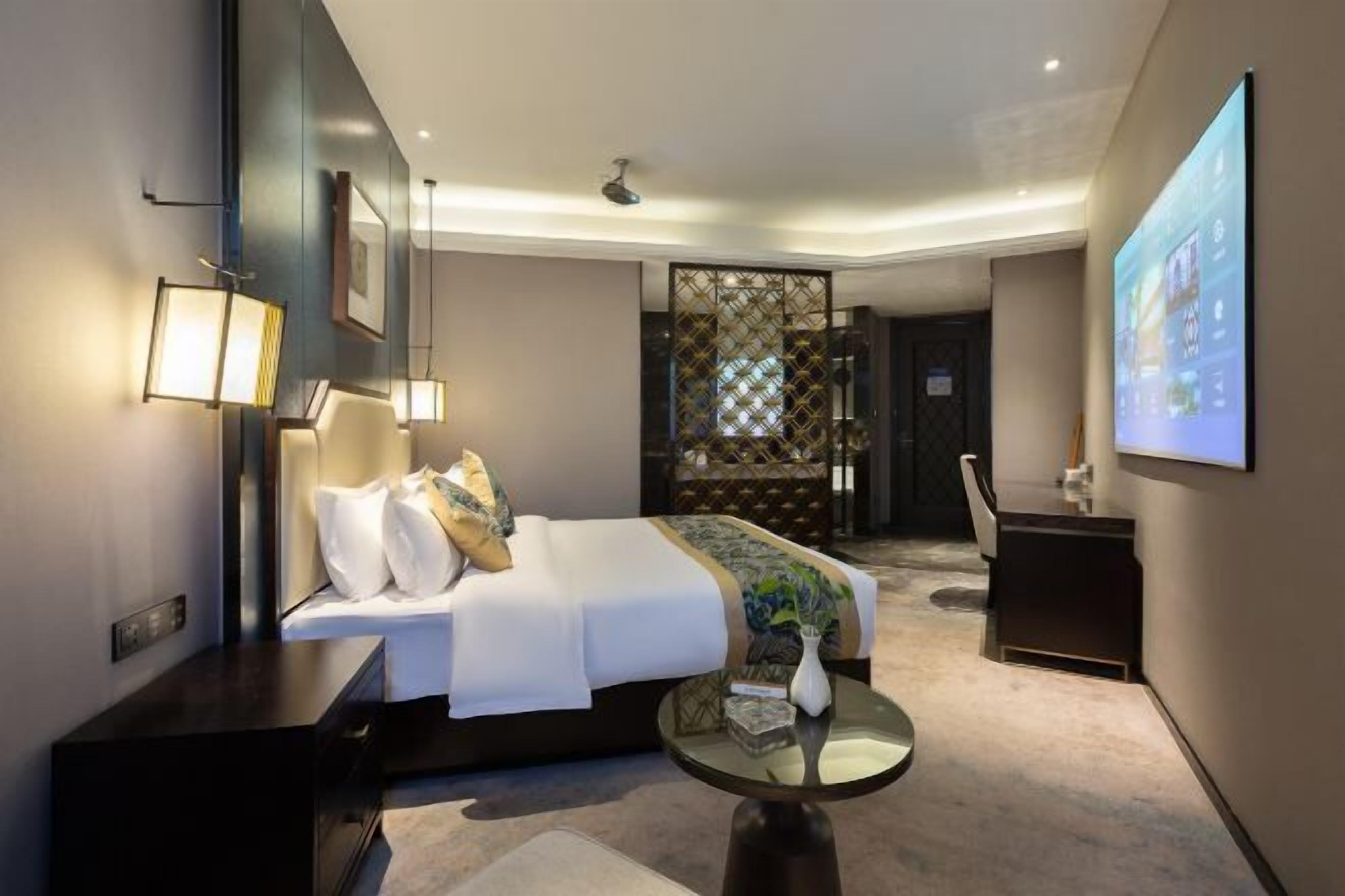 A modern hotel room with a bed, bedside table, lamp, and a screen displaying a cityscape.