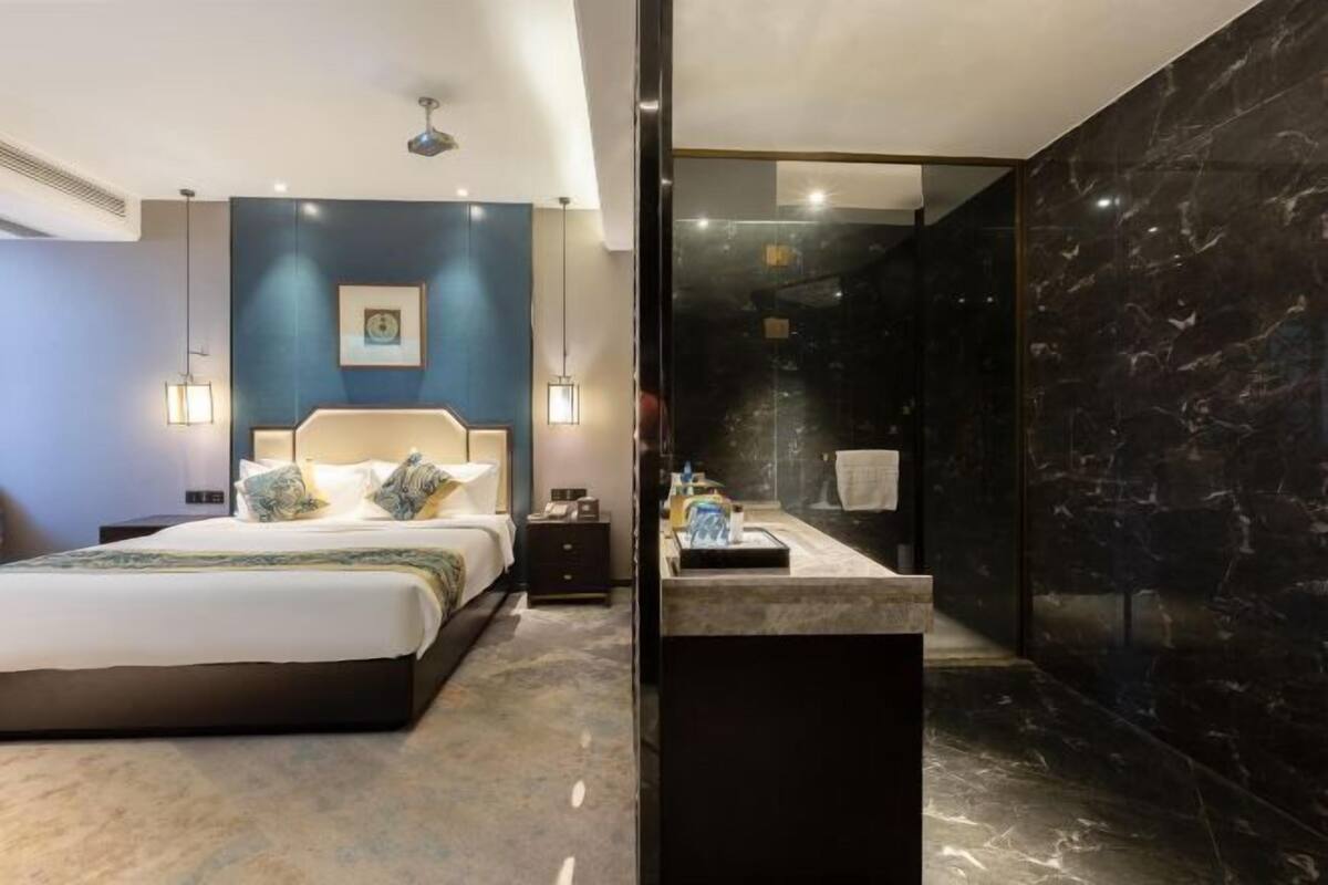 Une chambre d’hôtel moderne avec un lit, des lampes de chevet, une table de nuit, un tableau encadré au mur et une salle de bain dotée d’un plan de toilette et d’un lavabo en marbre.