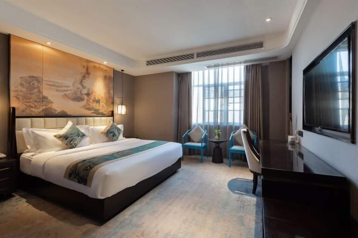 A hotel room with a large bed, a flat-screen TV, a desk, and a view of the city through large windows.