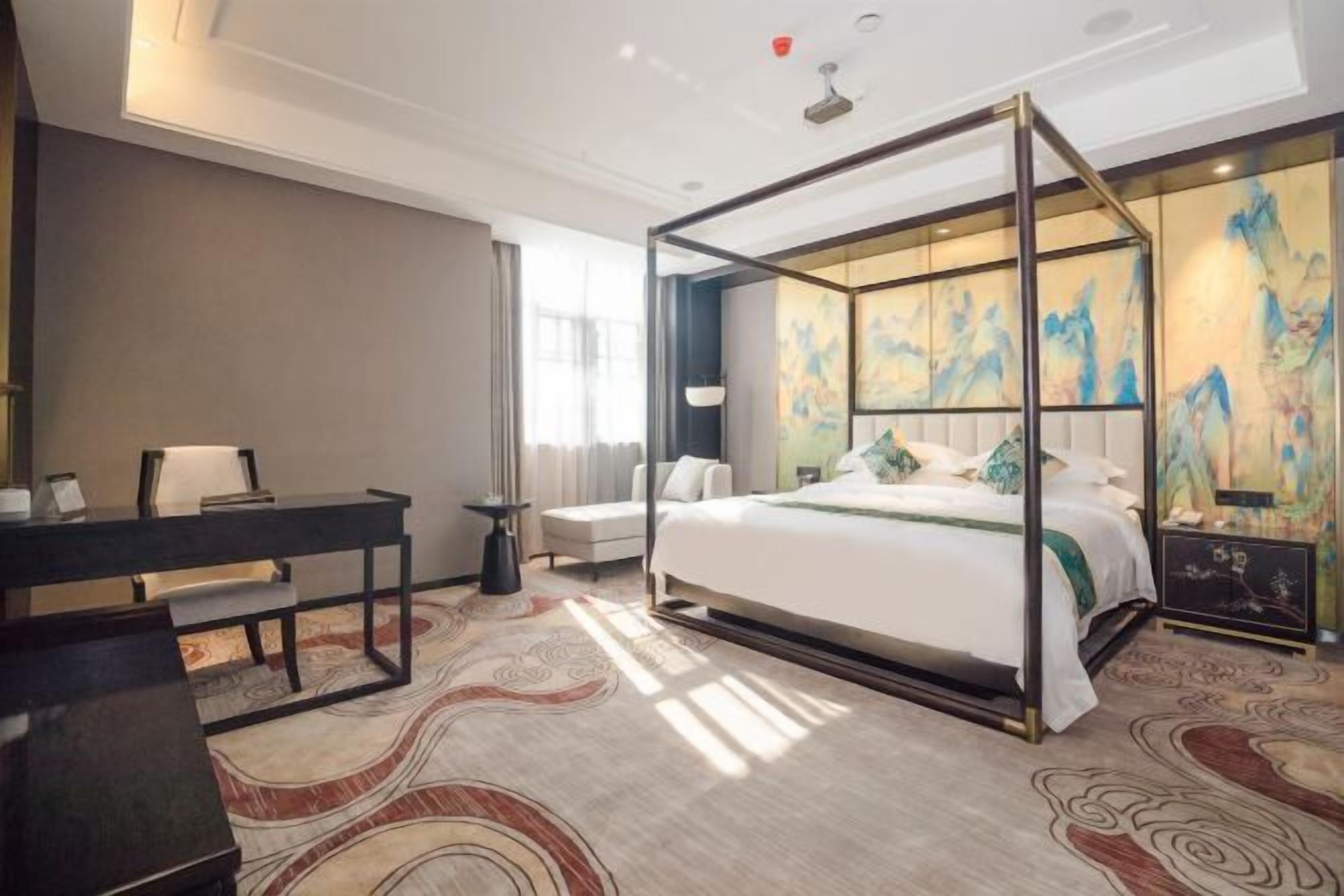 A bedroom with a four-poster bed, a desk, a chair, and a large painting on the wall.