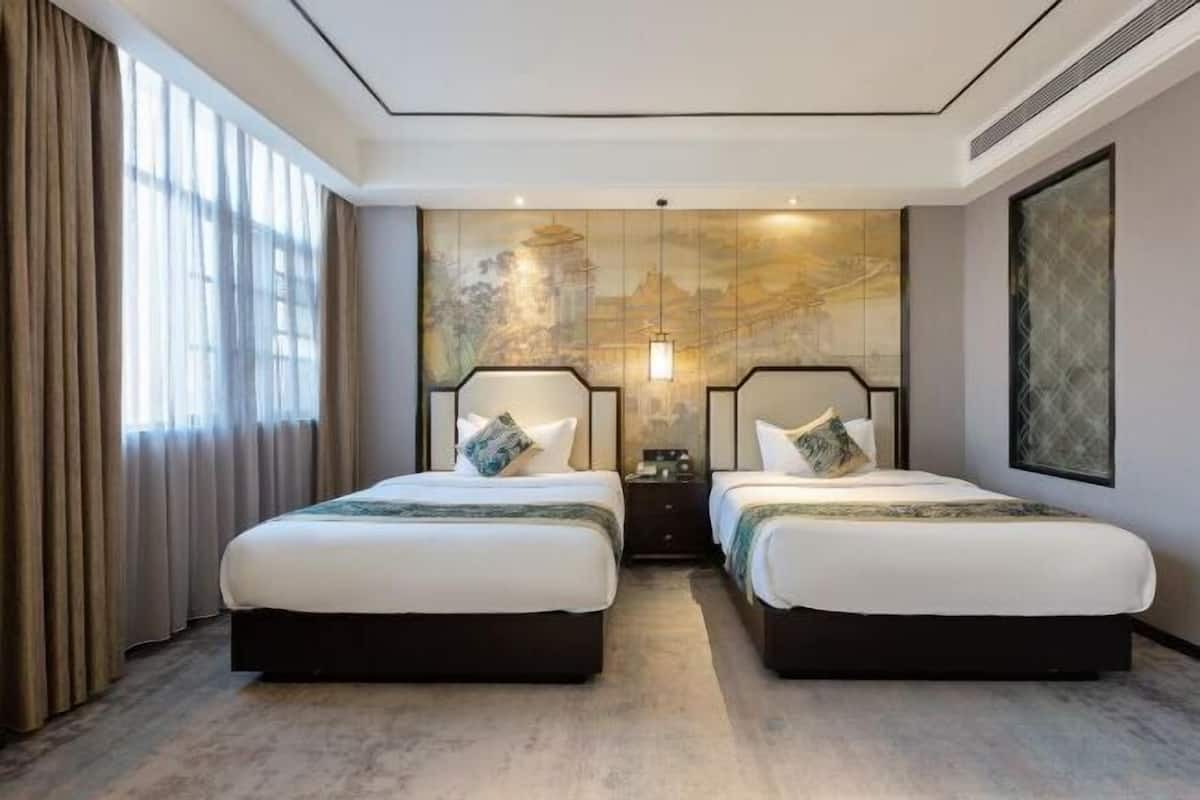 Une chambre d’hôtel avec deux lits, une grande fenêtre avec des rideaux, une œuvre d’art accrochée au mur et une table de chevet avec une lampe.