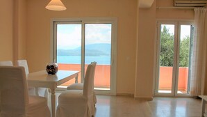 Una habitación con una mesa blanca, dos sillas blancas, un jarrón con flores y vistas a un cuerpo de agua y montañas a través de ventanas amplias.