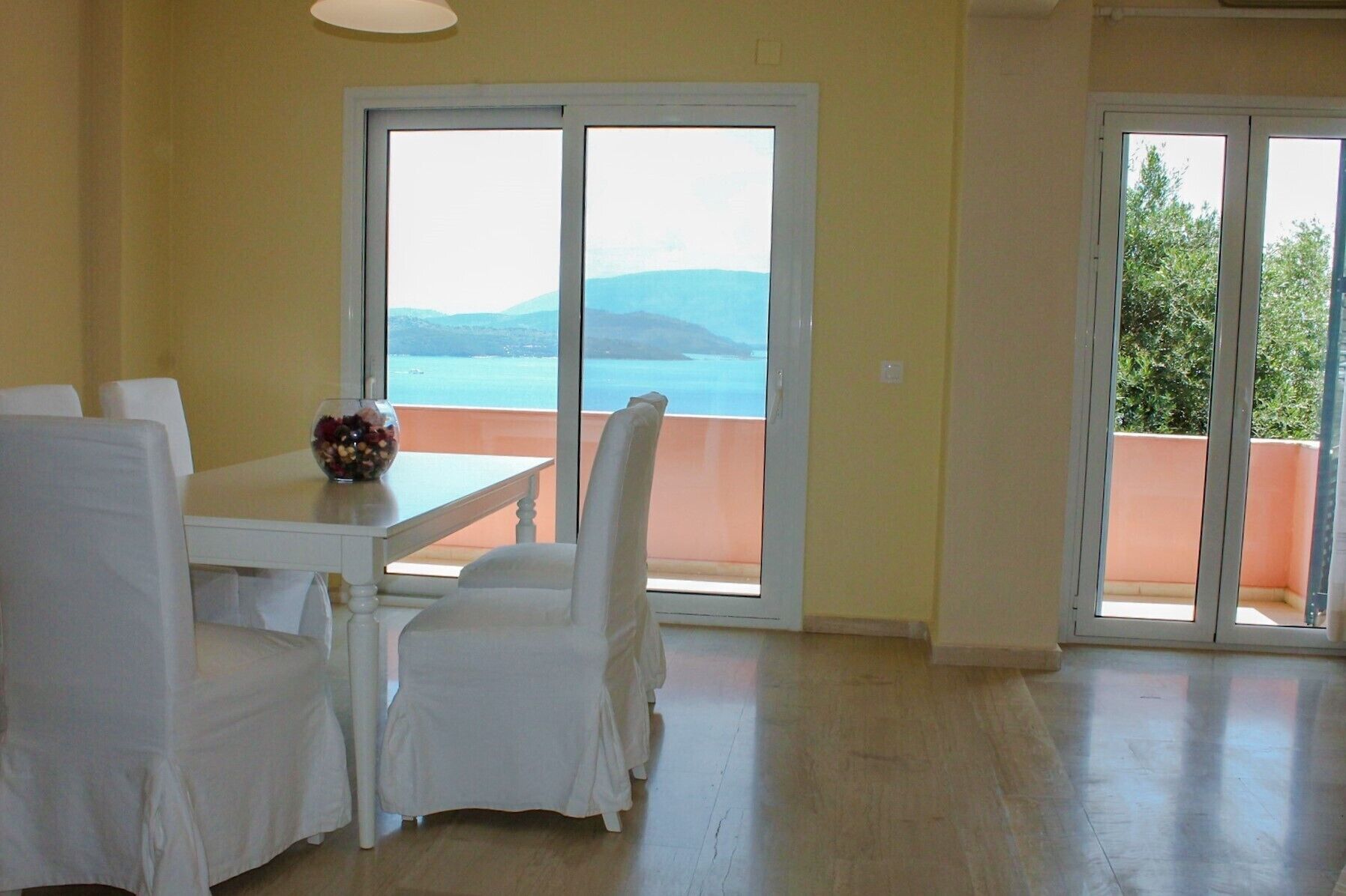 Uma área de refeições com mesa e cadeiras, uma tigela de frutas e grandes portas de vidro deslizantes que dão para uma varanda com vista para o mar e montanhas.