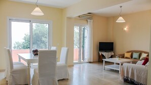 Amplia sala de estar con una mesa de comedor, sillas, sofá, televisión y un balcón con vista a árboles.