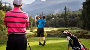 Een golfbaan met een bergachtige achtergrond, een golfer in de zwaai en een golftas op de grond.