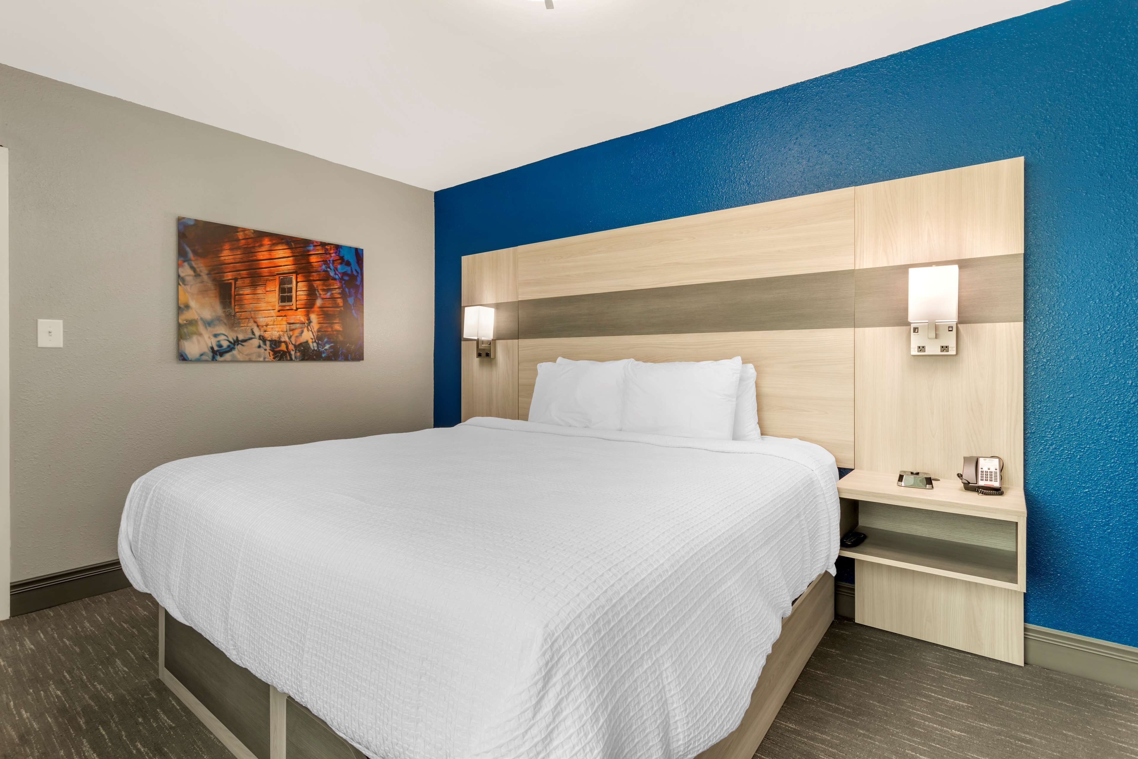 A hotel room with a large bed, a headboard with integrated lighting, a bedside table with a phone, and a painting on the wall.