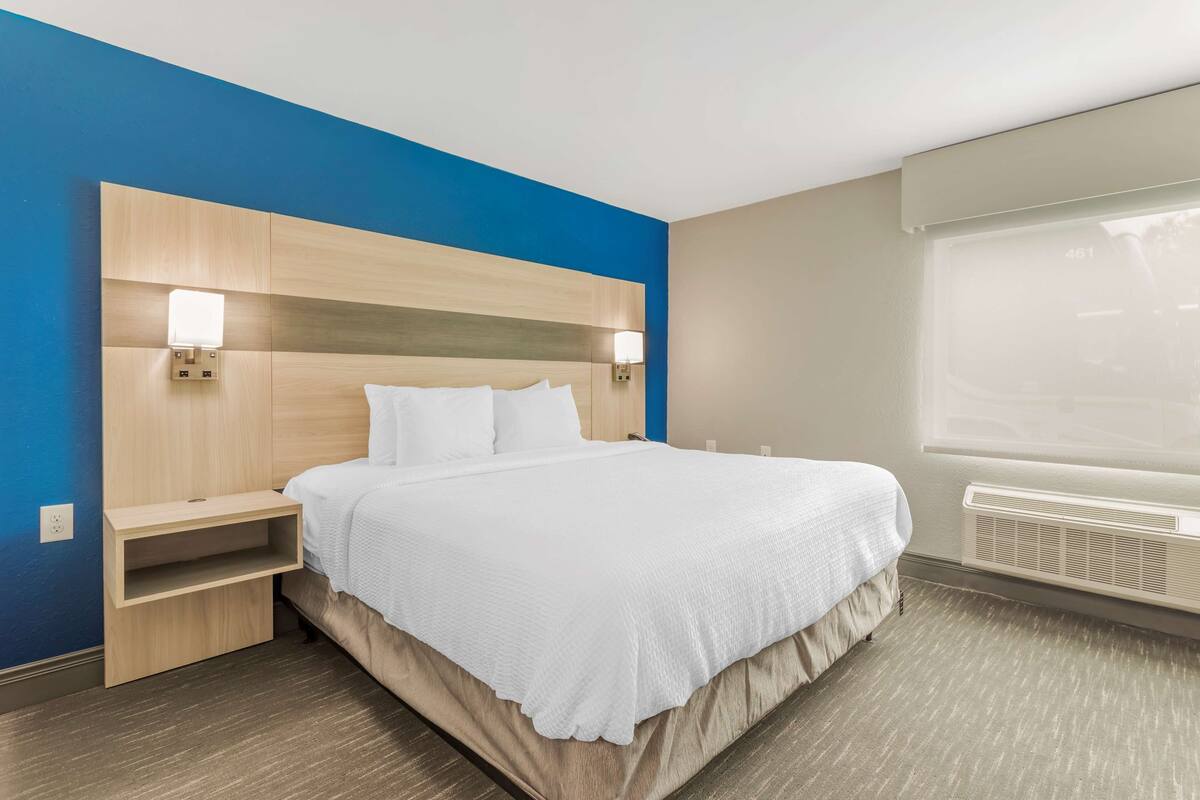 A hotel room with a bed, bedside tables, a window with blinds, and a wall-mounted air conditioning unit.