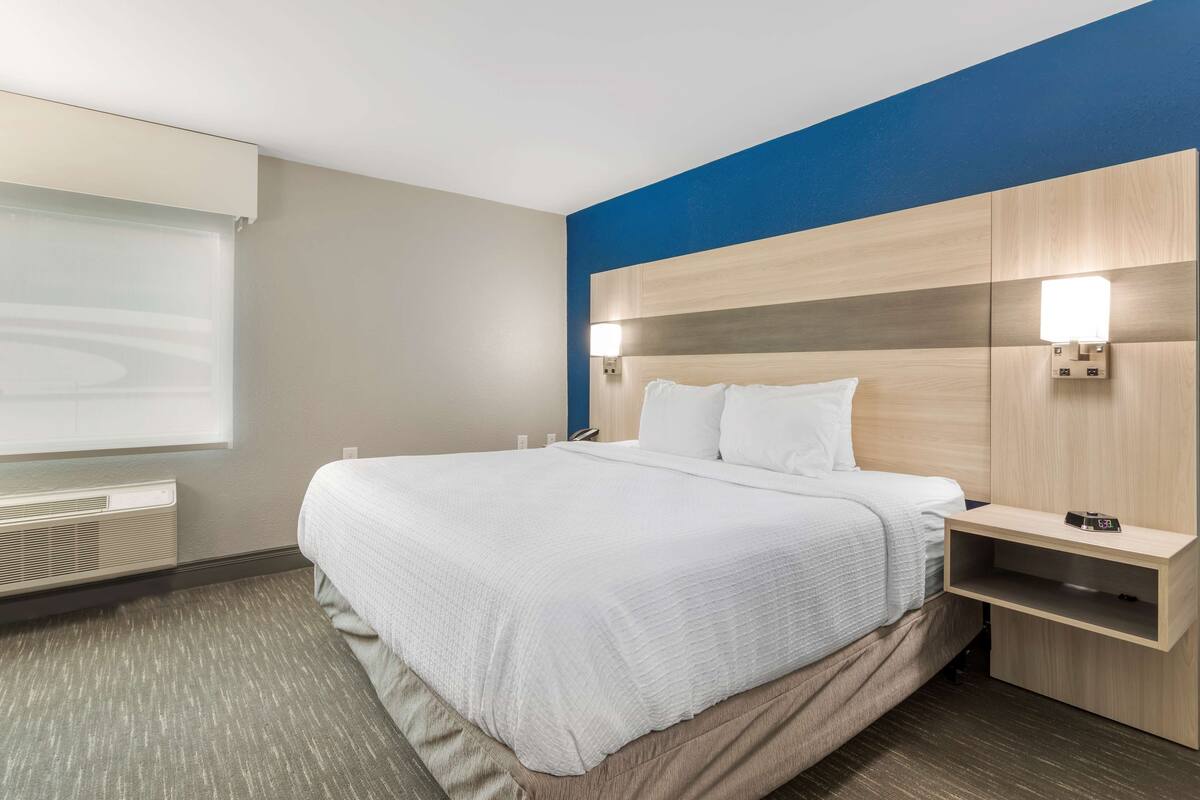 A hotel room with a large bed, a nightstand, a wall-mounted lamp, and a window with blinds.