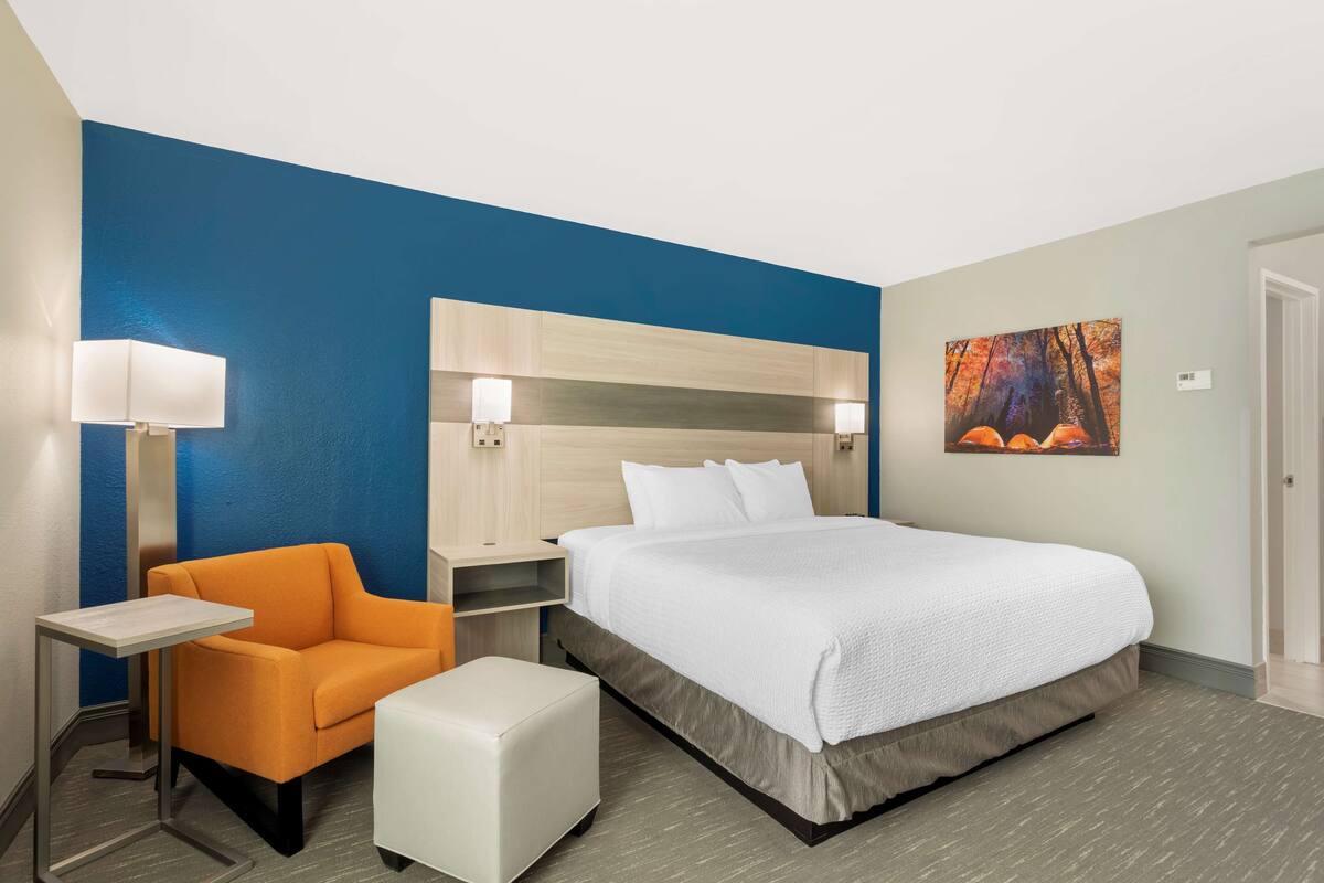 A hotel room with a bed, a nightstand, a chair, a small table, and a painting on the wall.