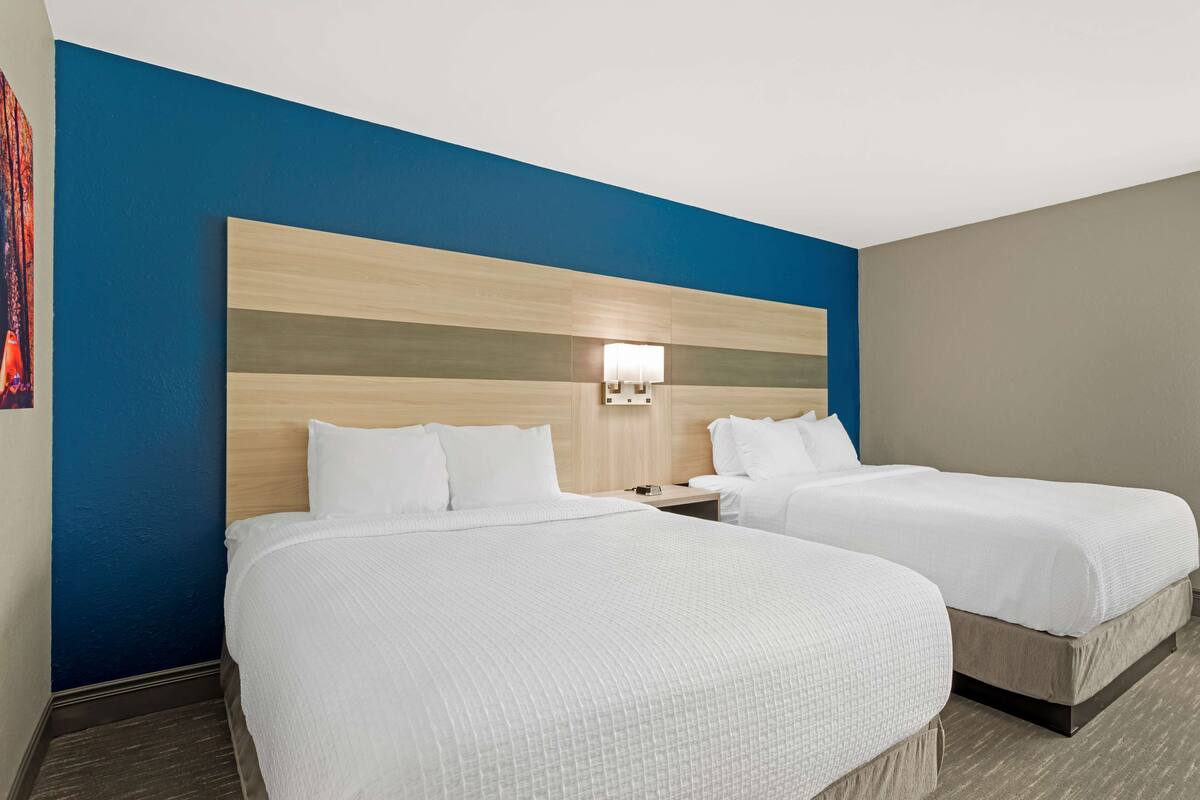 A hotel room with two beds, a wooden headboard, a nightstand, and a wall-mounted lamp.