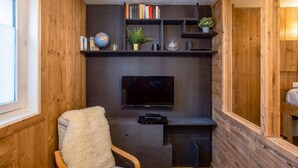 Una habitación acogedora con paredes de madera, un televisor de pantalla plana montado en una pared oscura, una estantería con libros y objetos decorativos, un sillón mullido y una pequeña mesa con un globo terráqueo y una planta.