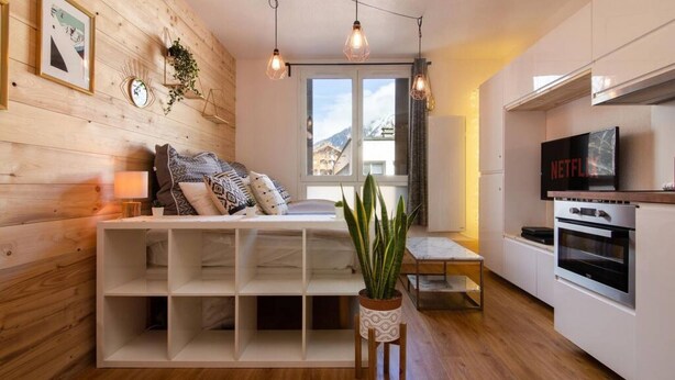 Una sala moderna con pared de madera, un sofá con cojines estampados, una mesa consola blanca, una planta en maceta y una zona de cocina con televisor y horno.