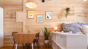 Una habitación acogedora con una pared de madera, una mesa de comedor con sillas, un sofá blanco con cojines estampados y una planta pequeña sobre una mesita auxiliar.