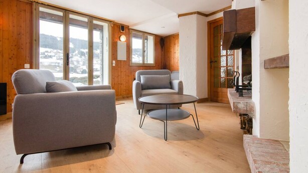 A modern living room with a sofa, two armchairs, and a round coffee table. Large windows offer a view of a snowy landscape.