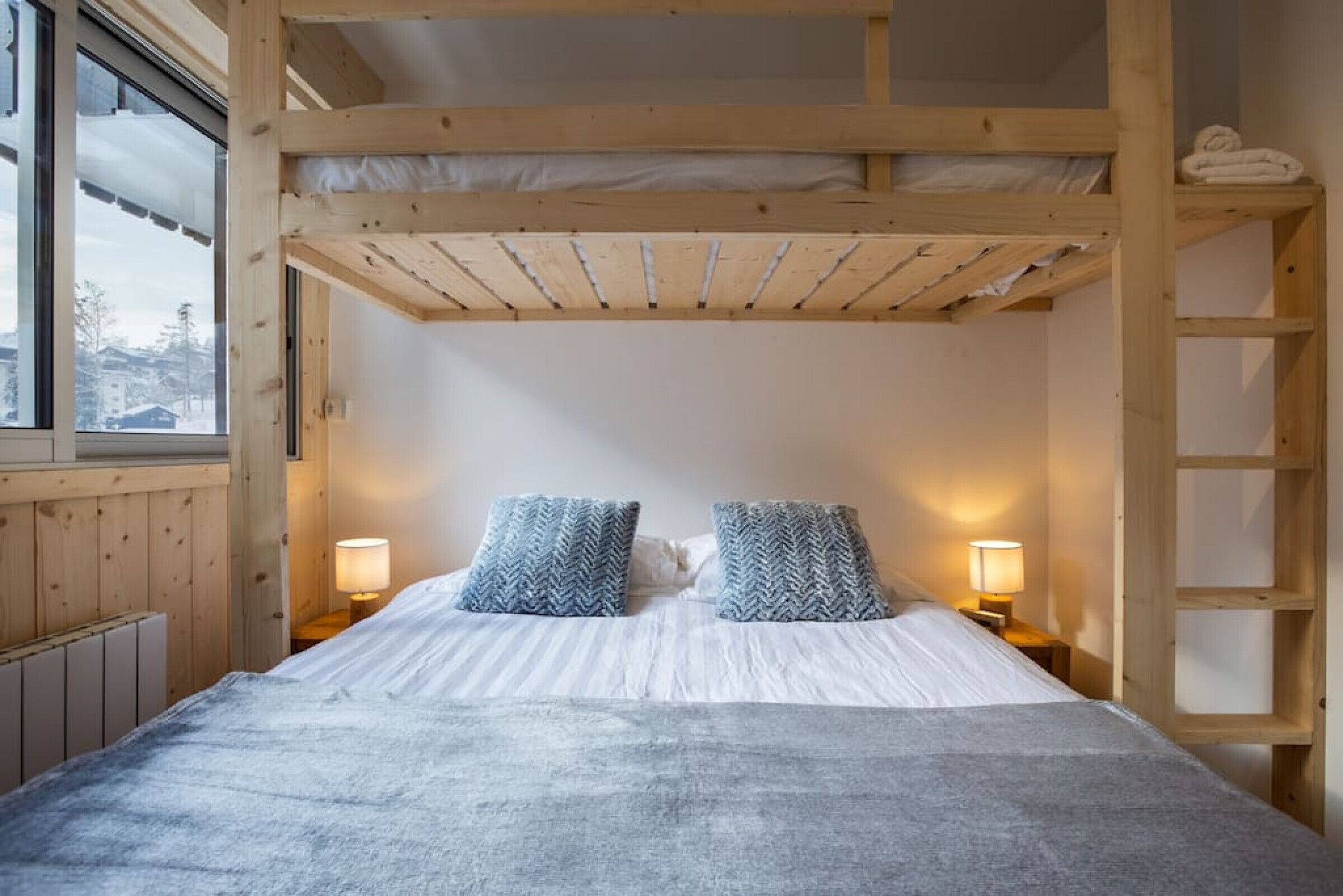 A bunk bed room with a wooden ceiling, two bedside lamps, and a view of the outside through a window.