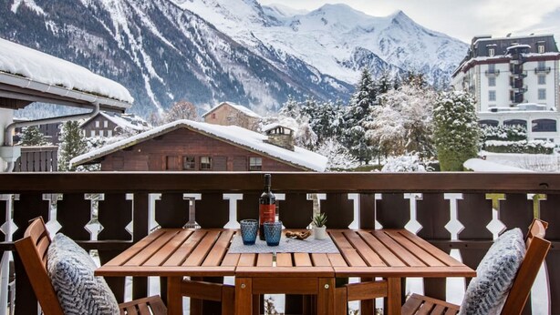 A wooden deck with a table and chairs, overlooking a snow-covered mountain range.