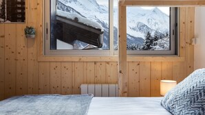 A cozy wooden room with a bed, a window with a snowy mountain view, and a small plant on the wall.
