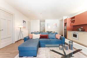 A modern living room with a sectional sofa, a glass coffee table, and a kitchen with a stove and cabinets.