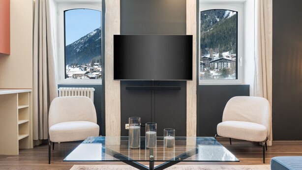 A modern living room with a large flat-screen TV mounted on a dark wall, two comfortable chairs, a glass coffee table, and a view of snow-covered mountains through large windows. - Montcler - Moderne&Spacieux 3CH Apart Hypercentre | Vue 180° (Chamonix-Mont-Blanc)