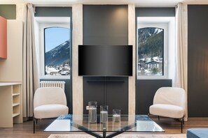 A modern living room with a large flat-screen TV mounted on a dark wall, two comfortable chairs, a glass coffee table, and a view of snow-covered mountains through large windows. - Montcler - Moderne&Spacieux 3CH Apart Hypercentre | Vue 180° (Chamonix-Mont-Blanc)