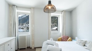 A bedroom with a bed, a chair, a radiator, and two windows with a view of a snowy village and mountains. - Montcler - Moderne&Spacieux 3CH Apart Hypercentre | Vue 180° (Chamonix-Mont-Blanc)