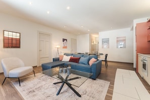 A modern living room with a blue sofa, a glass coffee table, and a white armchair. There is a dining area with a table and chairs in the background.