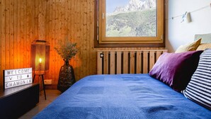 Une chambre avec un mur en bois, une fenêtre donnant sur la montagne, un lit avec une literie bleue, une table de chevet avec un petit panneau et une lampe.