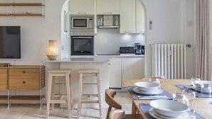 Una cocina moderna con gabinetes blancos, un microondas, un horno y una cafetera. Hay un comedor con una mesa preparada para dos personas, dos taburetes de bar y una lámpara.