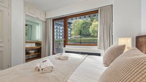 Habitación de hotel con una cama grande, ventana con vistas a edificios y árboles, tocador con espejo y mesita de noche.