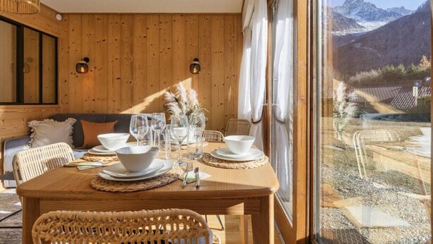A wooden dining table set with white plates, bowls, and wine glasses, with a view of mountains through a large window. - Modern Apart | MtBlanc View | 5mn Slopes | Parking (Chamonix-Mont-Blanc)