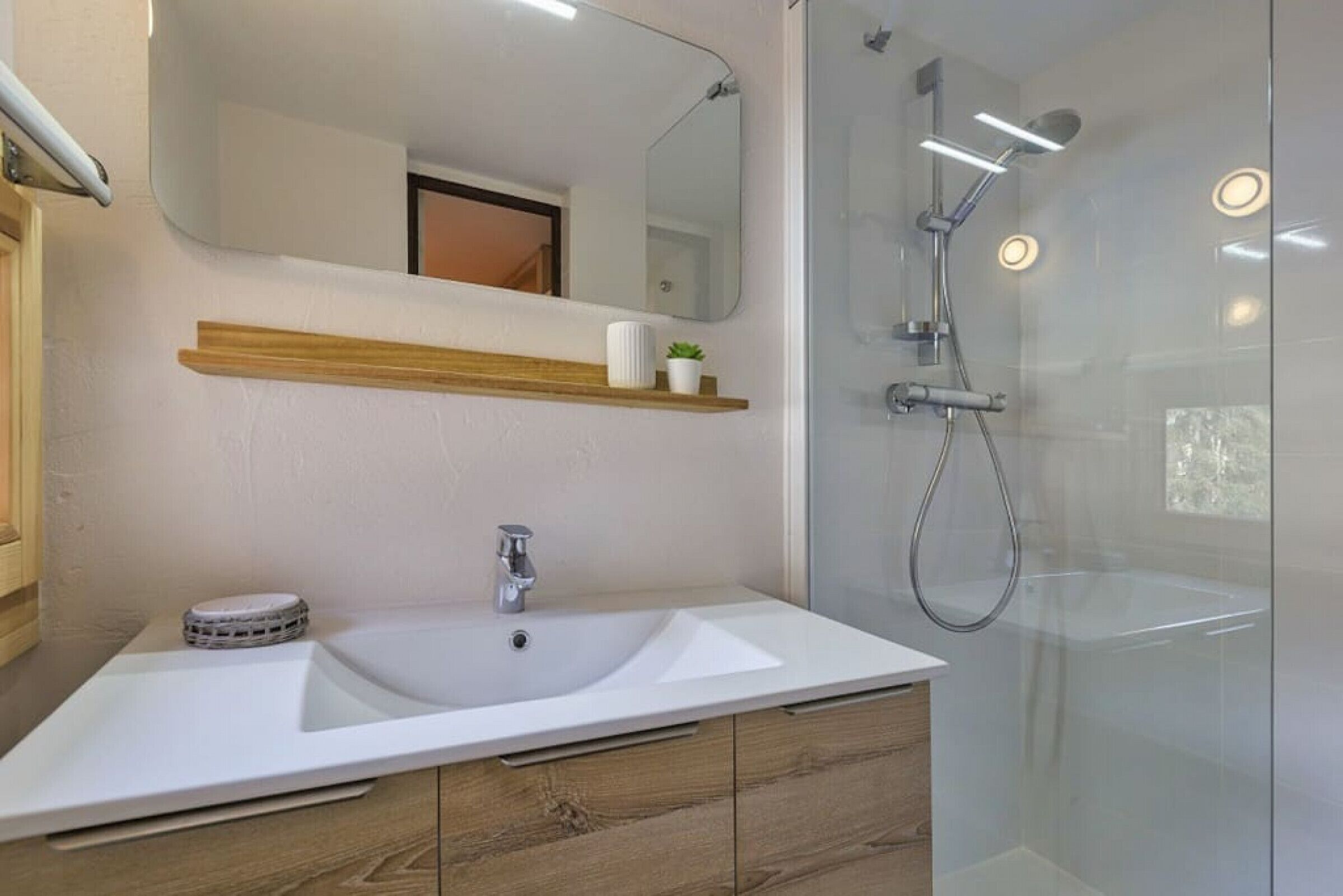 Une salle de bain moderne avec un lavabo blanc, des meubles en bois, un miroir et une douche à parois de verre.